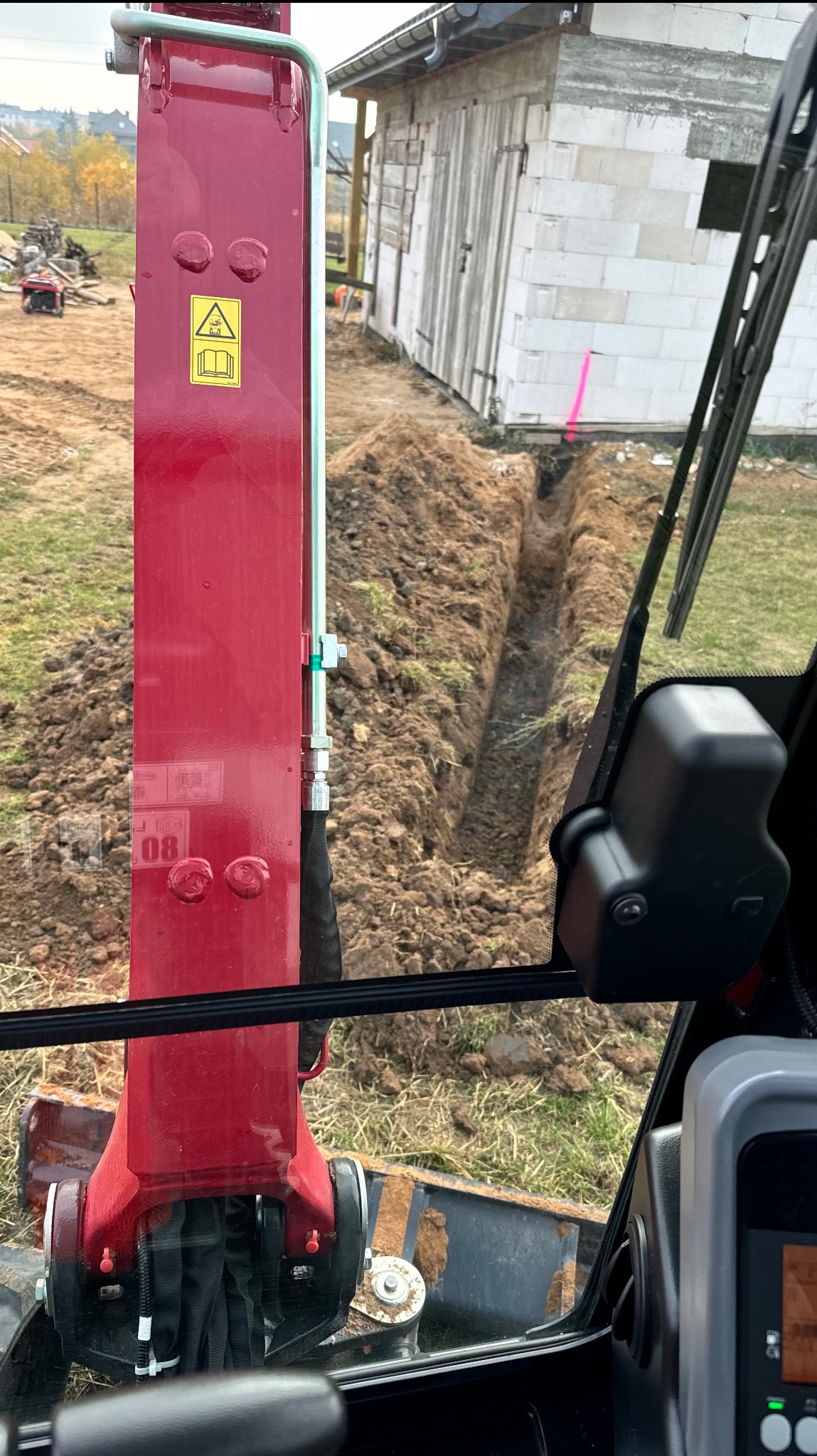 Widok z kabiny czerwonej minikoparki na wykopany wąski rów w ziemi, obok budynku z białych bloczków.
