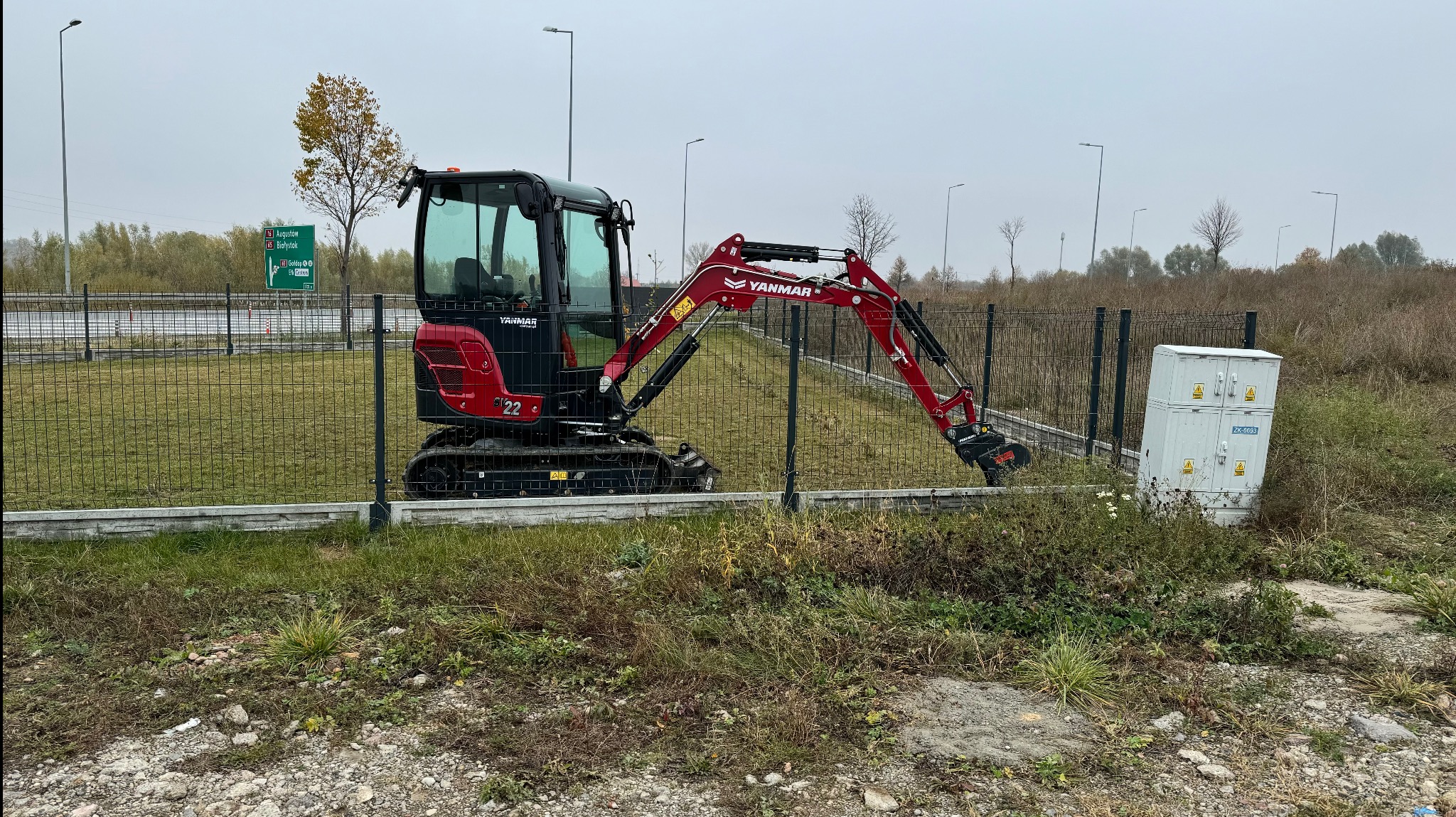 Czerwono-czarna minikoparka Yanmar 2.2 stoi na trawiastym terenie za metalowym ogrodzeniem, w tle widoczna droga ekspresowa i skrzynka elektryczna. Pochmurny dzień.