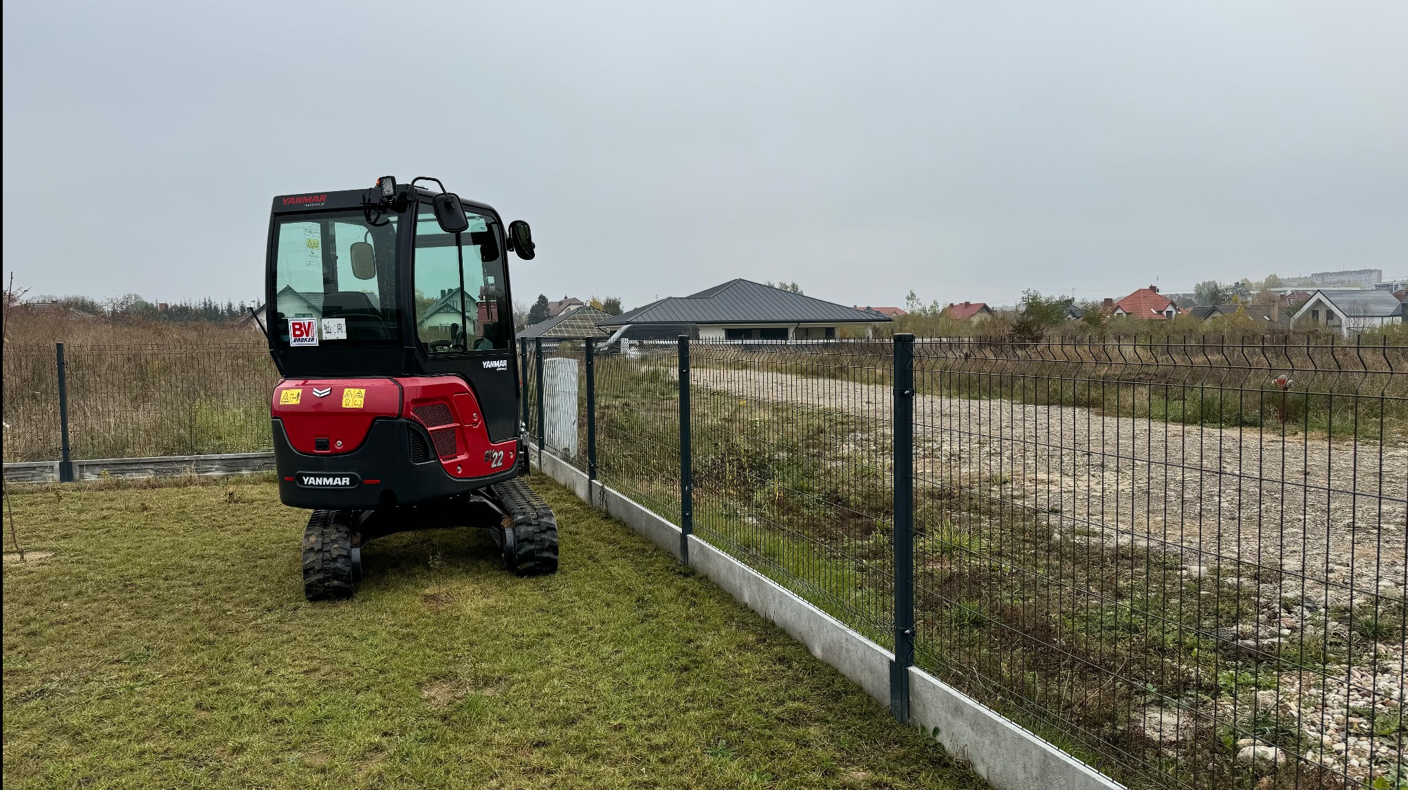 Czerwono-czarna minikoparka Yanmar stoi na trawie obok nowego ogrodzenia panelowego z podmurówką, widok na działkę z gruzem i domy w tle w pochmurny dzień.
