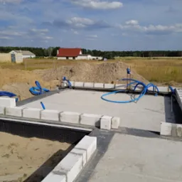 Fundamenty domu jednorodzinnego w trakcie budowy, z widoczną hydroizolacją i częściowo wymurowanymi ścianami z bloczków betonowych. W tle krajobraz z domami.