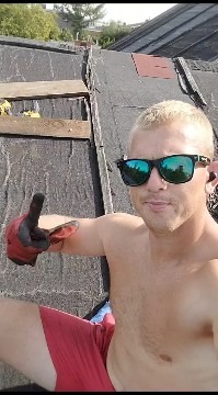 Mężczyzna w okularach i rękawicy na dachu krytym papą, w tle zielone drzewa i budynki. Nietypowy kadr z budowy dachu.