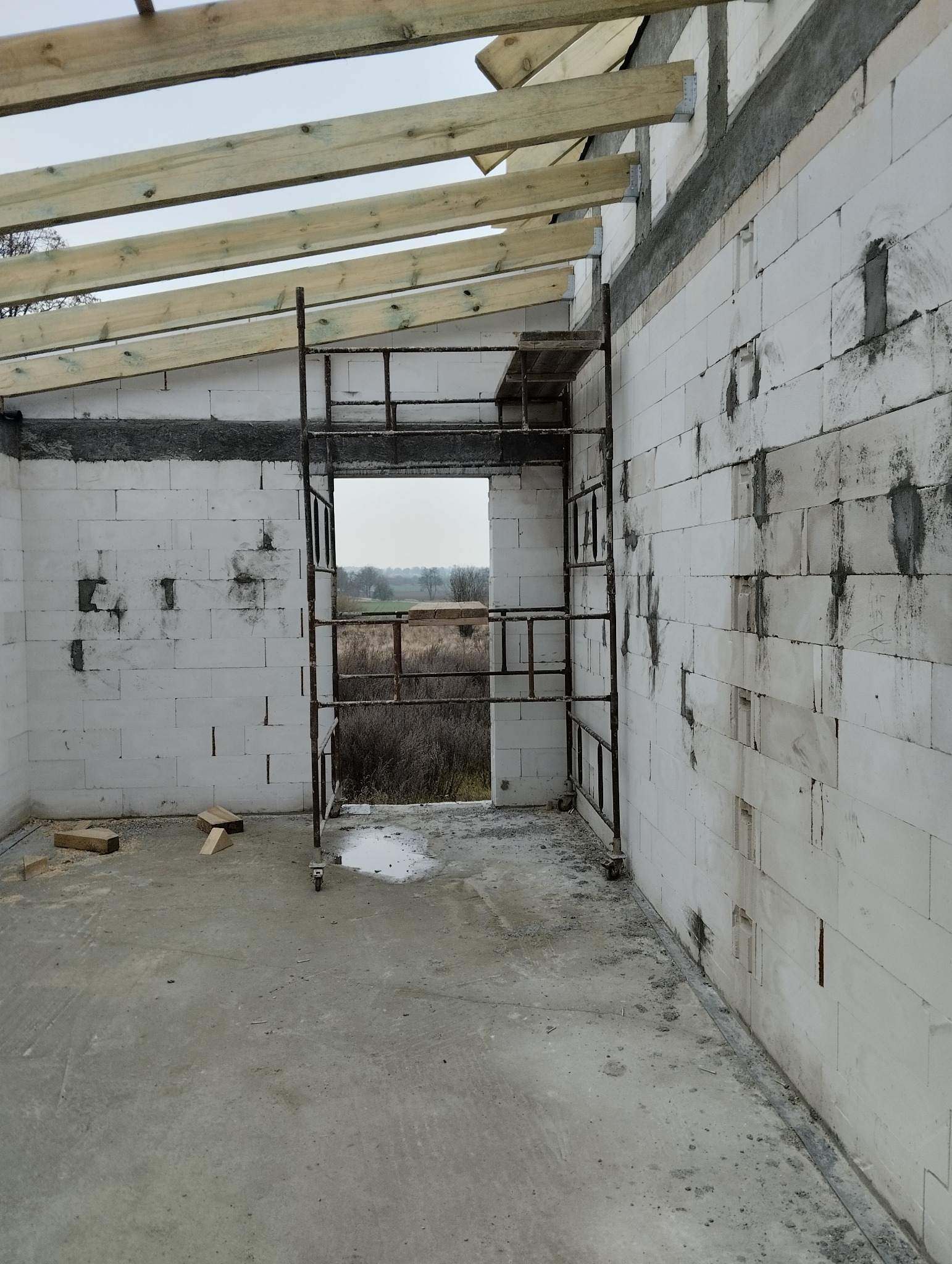Wnętrze surowego domu w trakcie budowy: białe ściany z betonu komórkowego, drewniane belki stropowe i metalowe rusztowanie przy otworze okiennym.