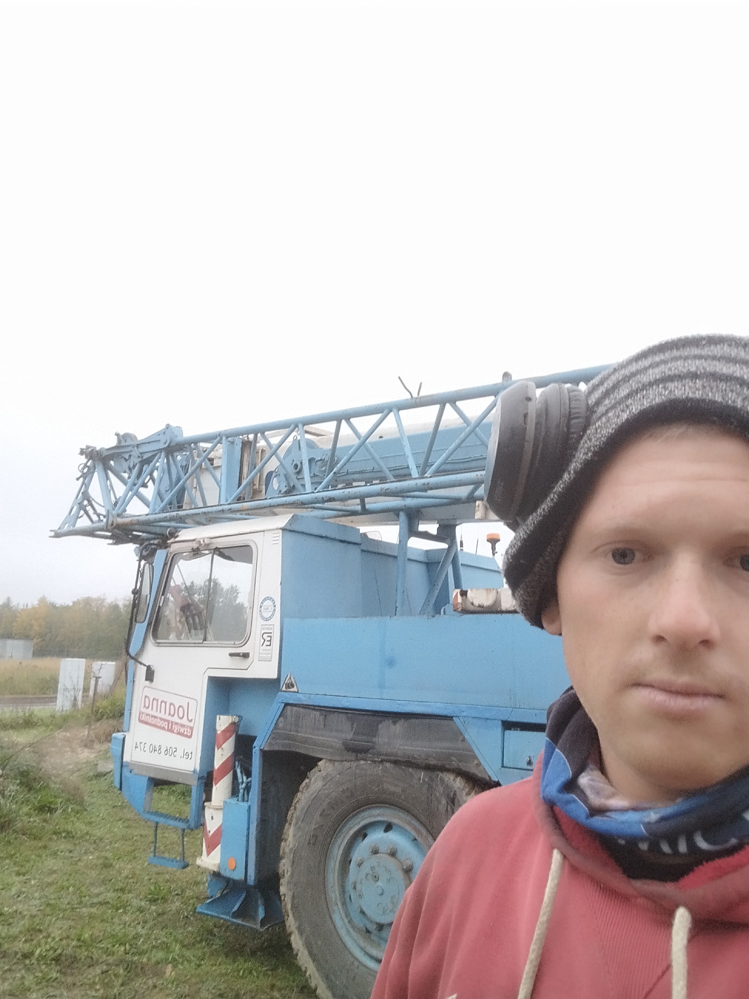 Mężczyzna w czapce i słuchawkach pozuje na tle niebiesko-białego dźwigu samochodowego z napisem 'Dandpol' na boku, stojącego na trawiastym terenie.