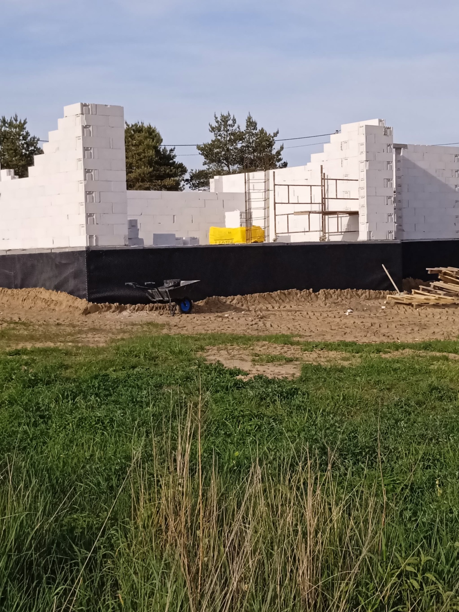 Budowa domu z bloczków w Sośnie. Widoczne fundamenty z izolacją, częściowo wymurowane ściany, rusztowanie i taczka. Prace budowlane w toku.