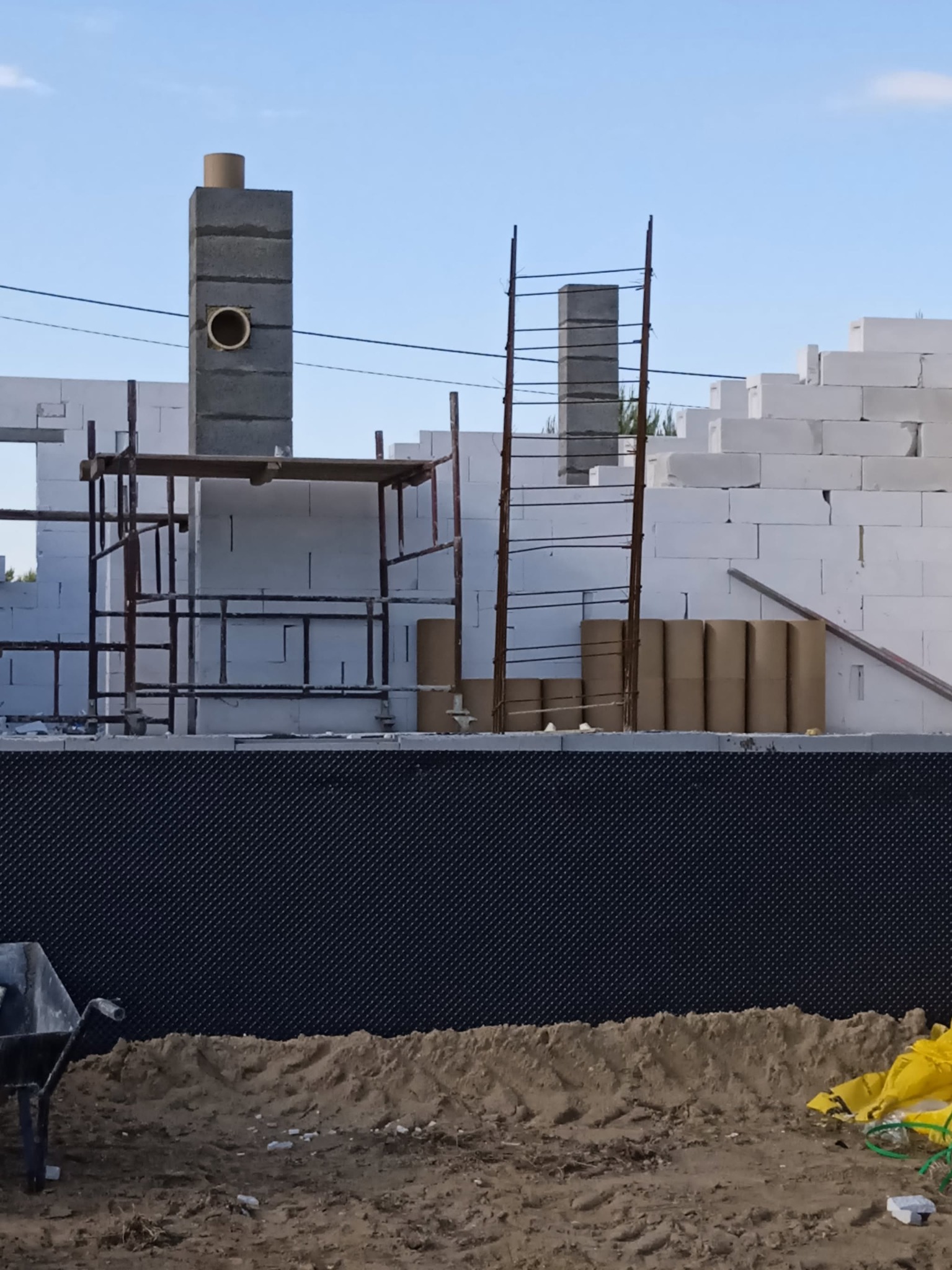 Wznoszenie murów z białych bloczków na fundamencie. Widoczne rusztowania, komin w budowie i materiały izolacyjne. Etap budowy domu w słoneczny dzień.