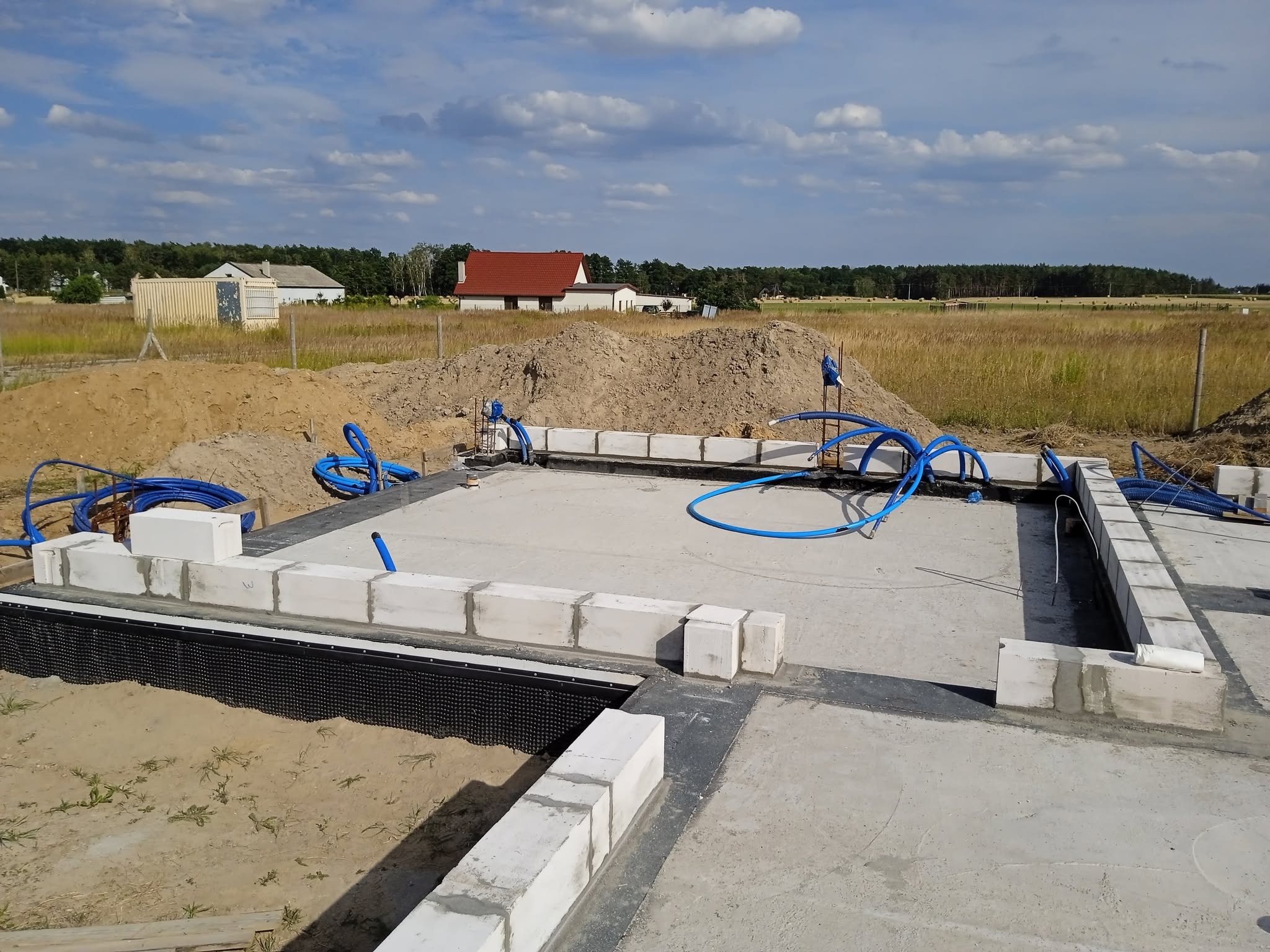 Fundamenty domu jednorodzinnego w trakcie budowy, z widoczną hydroizolacją i częściowo wymurowanymi ścianami z bloczków betonowych. W tle krajobraz z domami.
