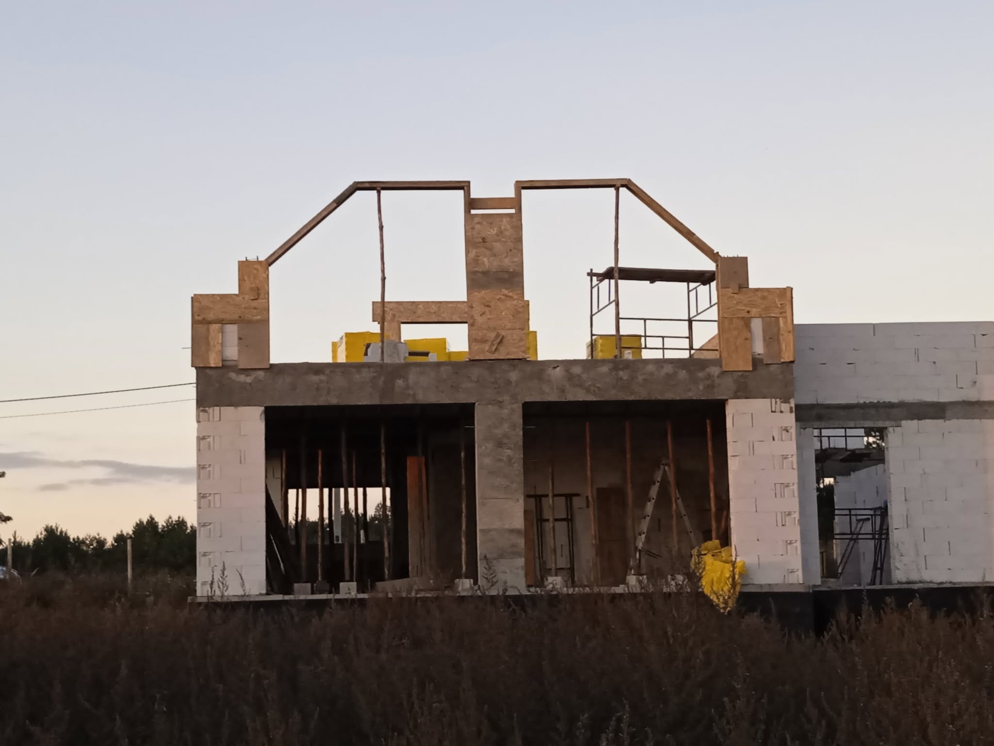 Niedokończony dom z białego pustaka, widoczne drewniane elementy konstrukcyjne dachu i rusztowanie. W tle zieleń drzew i niebo o zmierzchu.