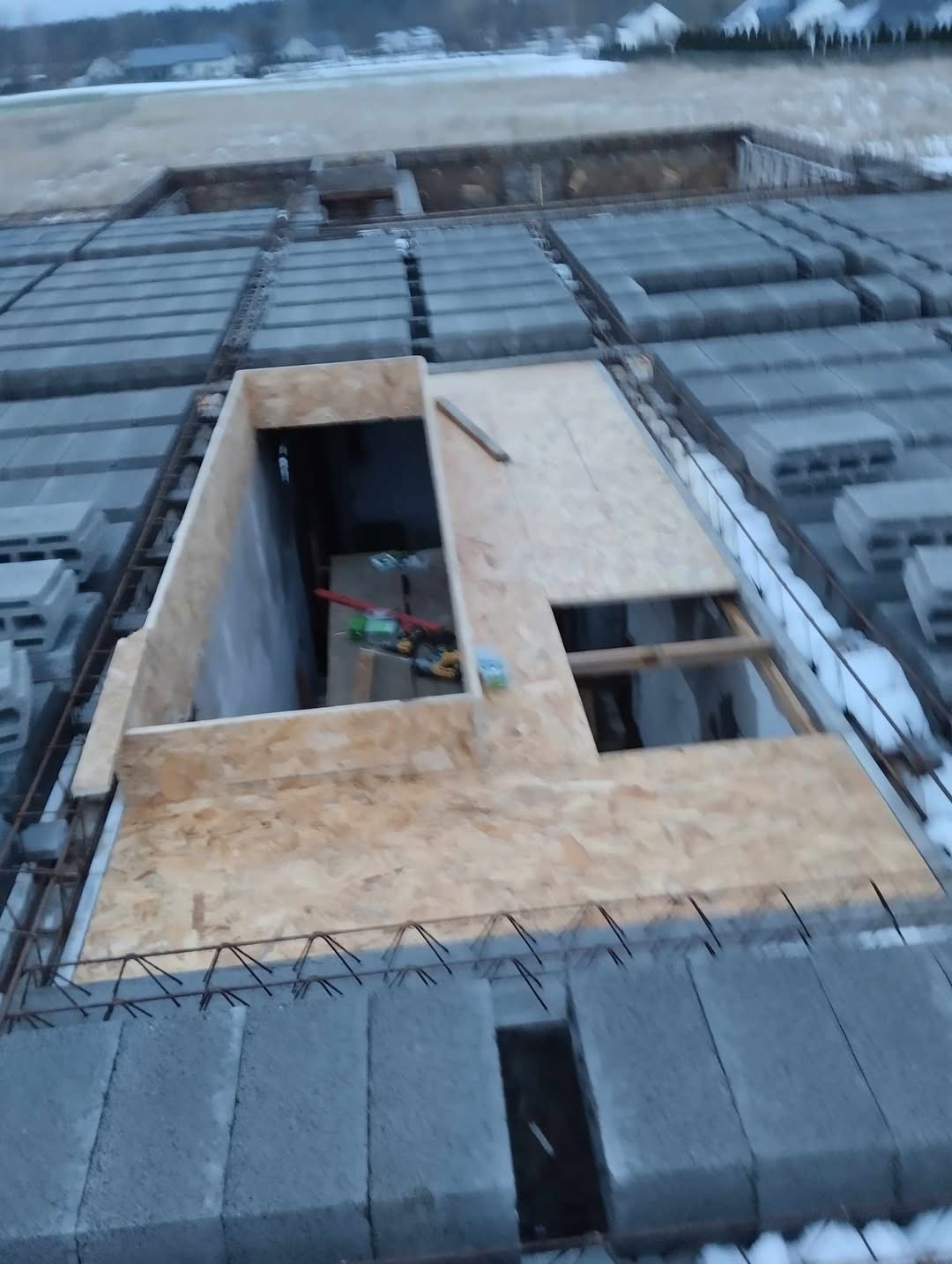 Wykopany fundament z częściowo ułożonymi bloczkami betonowymi i szalunkiem z desek. Widoczna stal zbrojeniowa. Zima w tle.