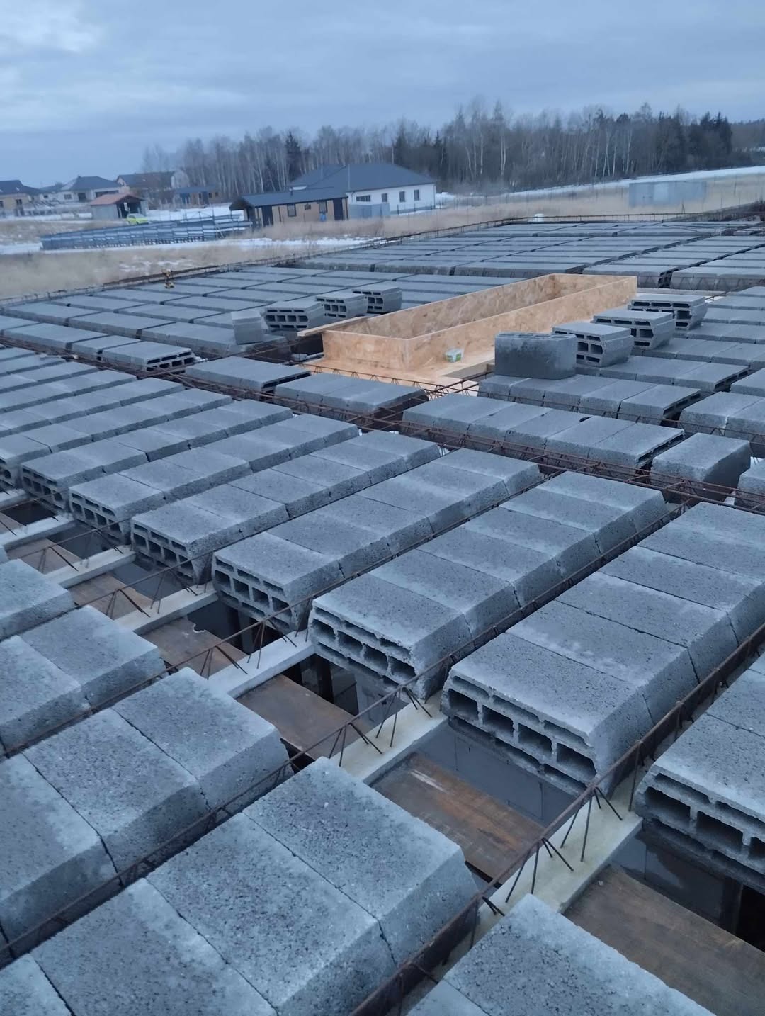 Szare bloczki betonowe ułożone na zbrojeniu, tworzące fundament pod budynek. Widoczna drewniana forma. Tło stanowią domy i drzewa pod pochmurnym niebem.