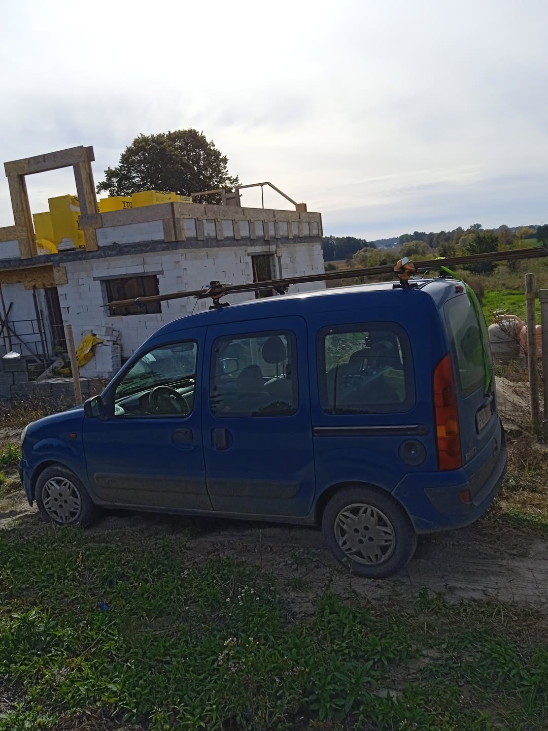 Niebieski samochód dostawczy na tle budowy domu z białego pustaka. Widoczne żółte materiały izolacyjne na dachu. Krajobraz wiejski, pochmurne niebo.