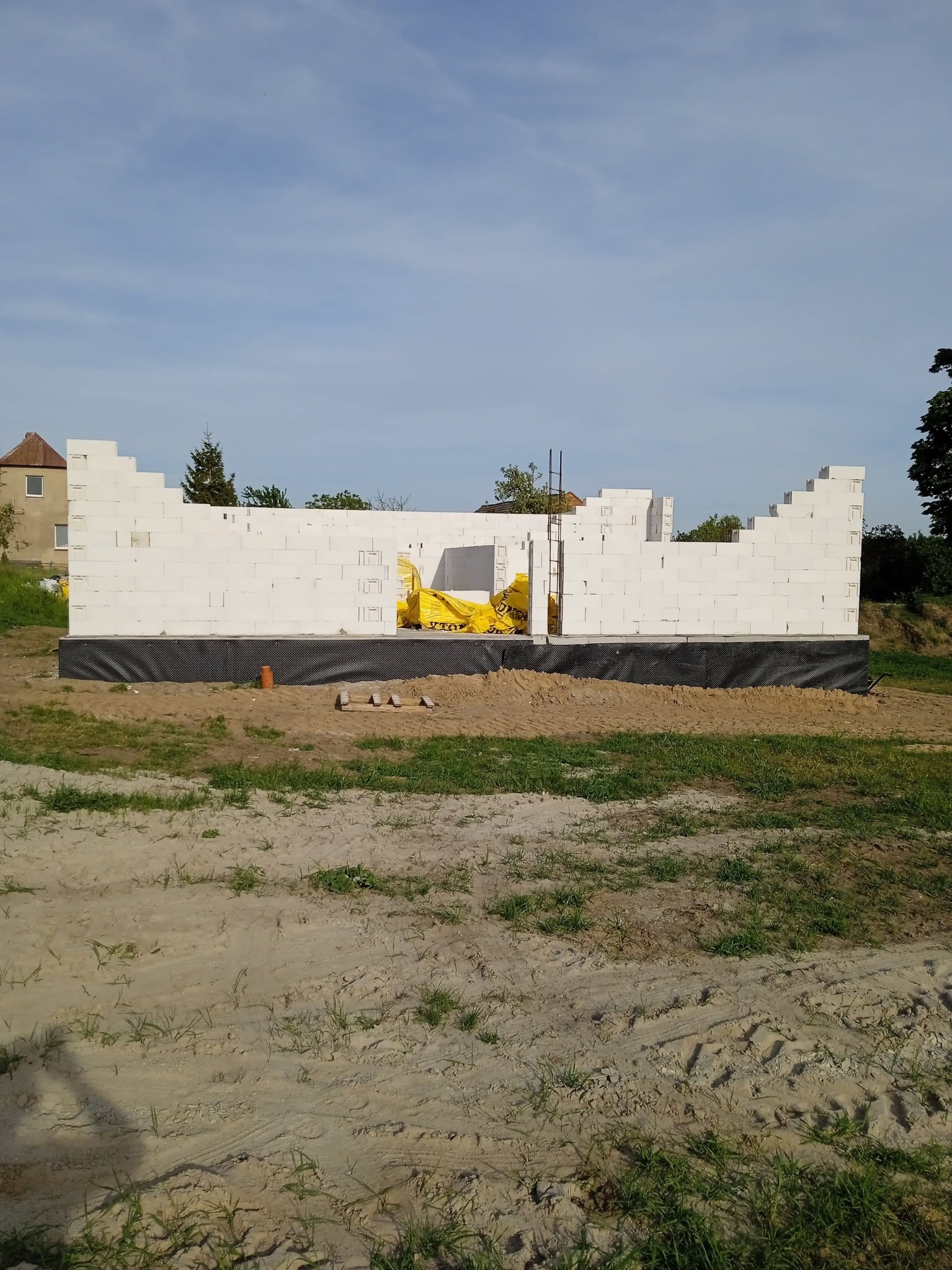 Wznoszenie murów domu z białych bloczków, widoczne żółte worki z materiałami budowlanymi wewnątrz konstrukcji. Budowa w toku, widok na niebo i fragment budynku w tle.
