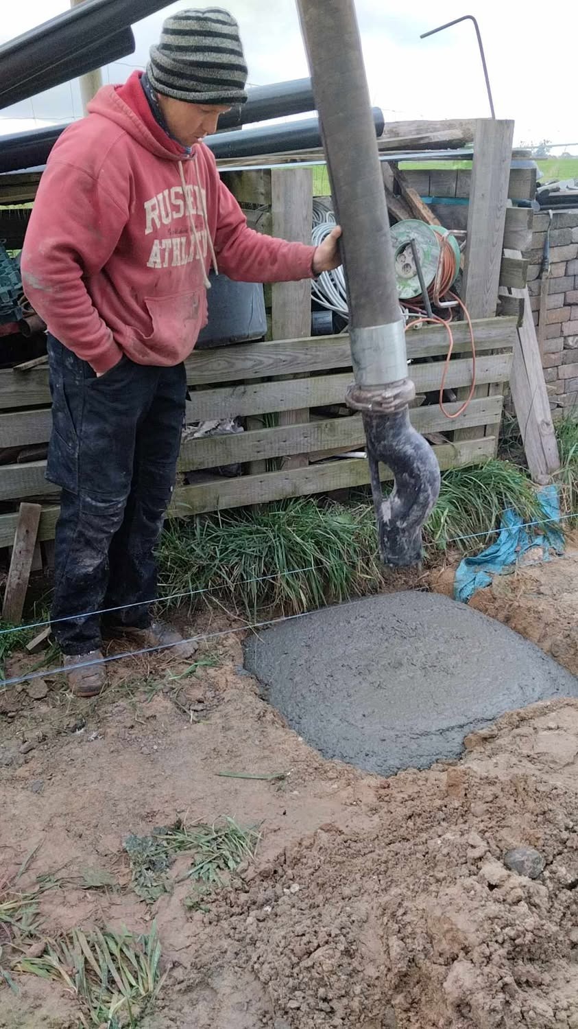 Wylewka betonu do fundamentu domu jednorodzinnego. Pracownik w czerwonej bluzie obsługuje pompę do betonu. Widoczne szalunki i świeży beton.