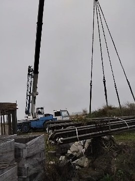 Żuraw przenosi stalowe belki na placu budowy. Widoczne elementy szalunku i palety z bloczkami. Pochmurne niebo w tle.