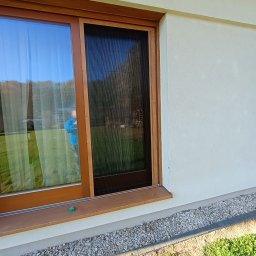 MiK Consulting Michał Kubiczek - Zewnętrzne okno PCV z moskitierą plisowaną, brązowa rama, widoczny refleks osoby w oknie, elewacja budynku w tle.