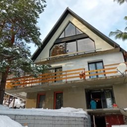 MiK Consulting Michał Kubiczek - Dom w stanie surowym, z zamontowanymi oknami o nietypowym, trójkątnym kształcie na poddaszu, drewniana balustrada balkonu, widoczne ślady prac budowlanych i zimowa sceneria.