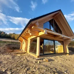 Nowoczesny dom z bali z dużymi, przesuwnymi oknami, jasne drewno, błękitne niebo, teren w trakcie zagospodarowania.