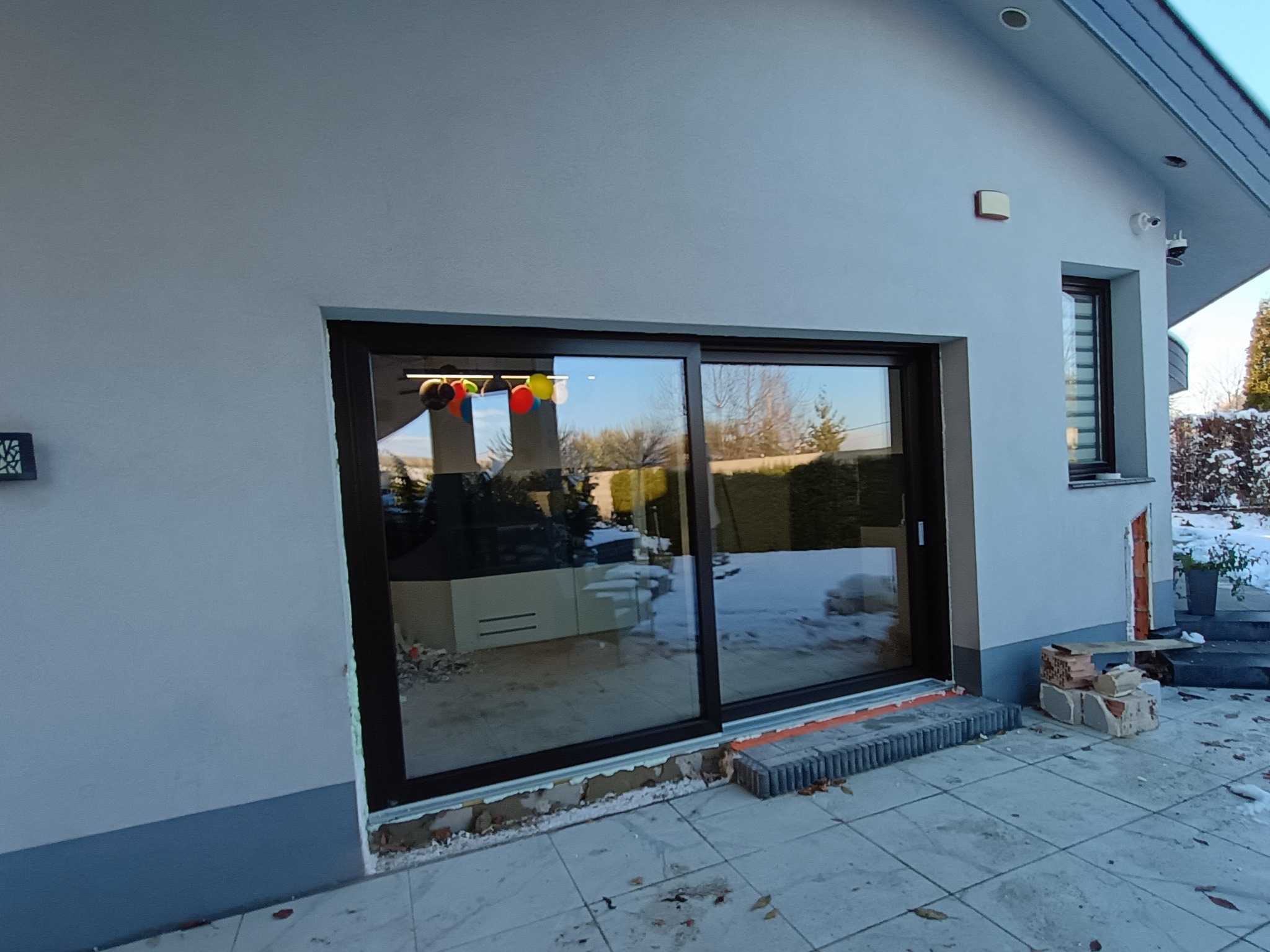 Montaż dużych, przesuwnych drzwi tarasowych z ciemną ramą w jasnym budynku, widoczne ślady prac montażowych na zewnątrz i balony wewnątrz.