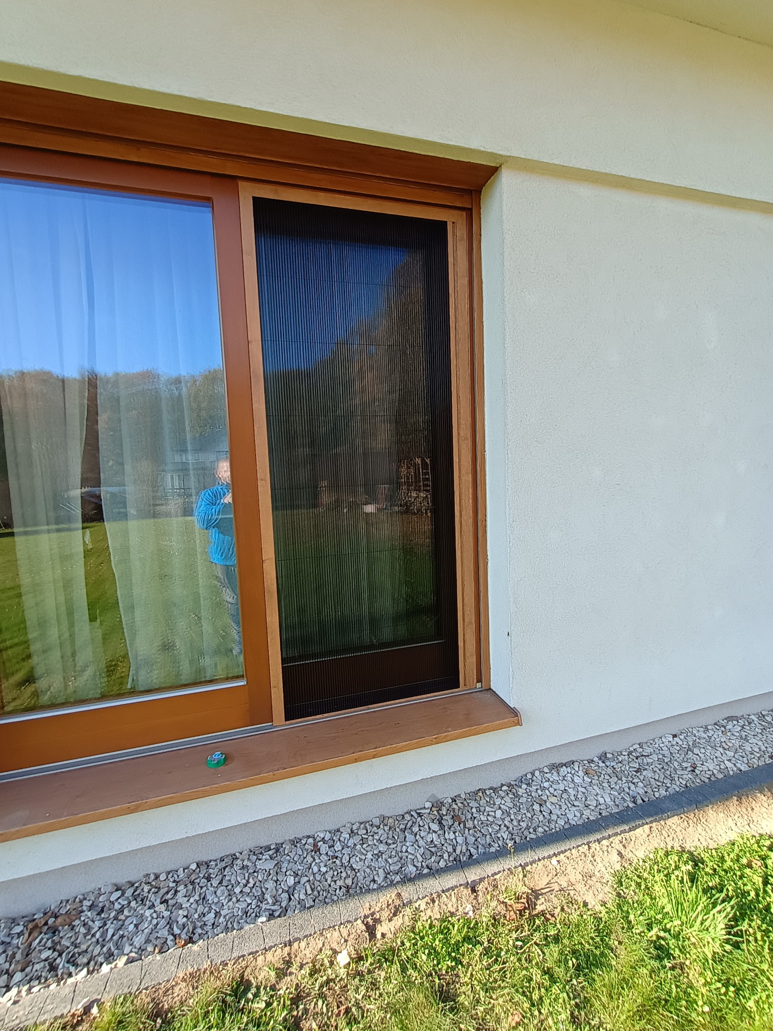 Zewnętrzne okno PCV z moskitierą plisowaną, brązowa rama, widoczny refleks osoby w oknie, elewacja budynku w tle.
