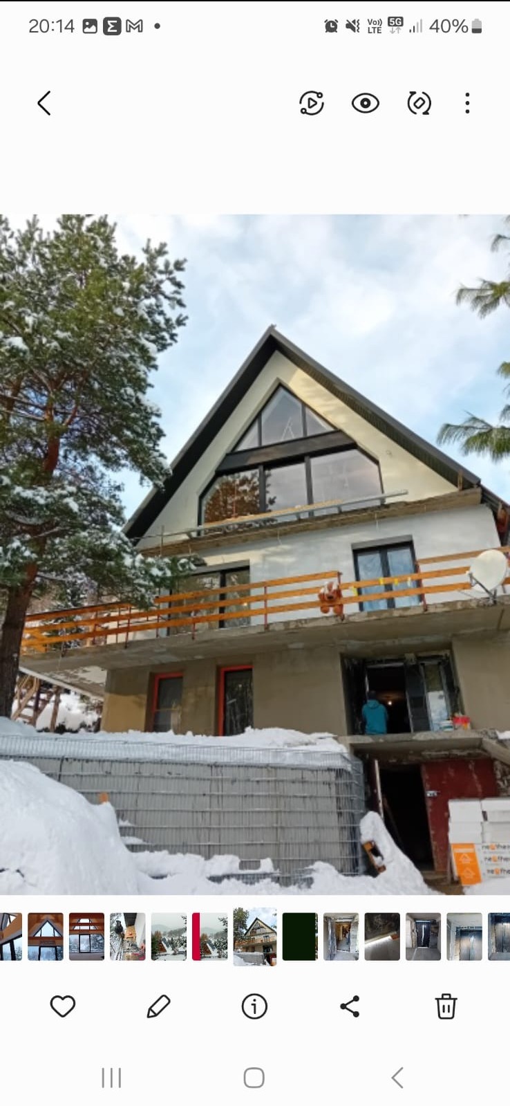 Dom w stanie surowym, z zamontowanymi oknami o nietypowym, trójkątnym kształcie na poddaszu, drewniana balustrada balkonu, widoczne ślady prac budowlanych i zimowa sceneria.