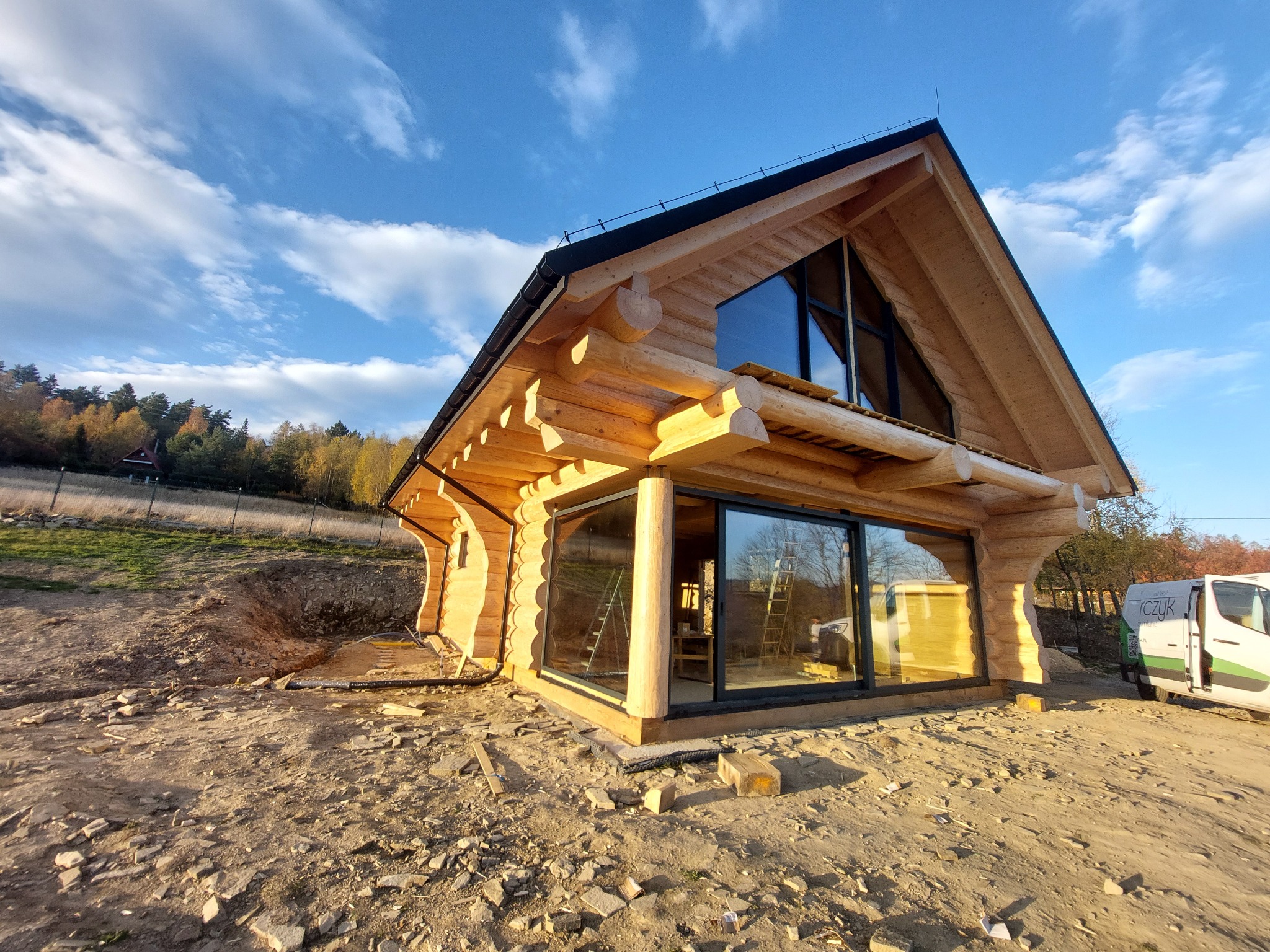 Nowoczesny dom z bali z dużymi, przesuwnymi oknami, jasne drewno, błękitne niebo, teren w trakcie zagospodarowania.