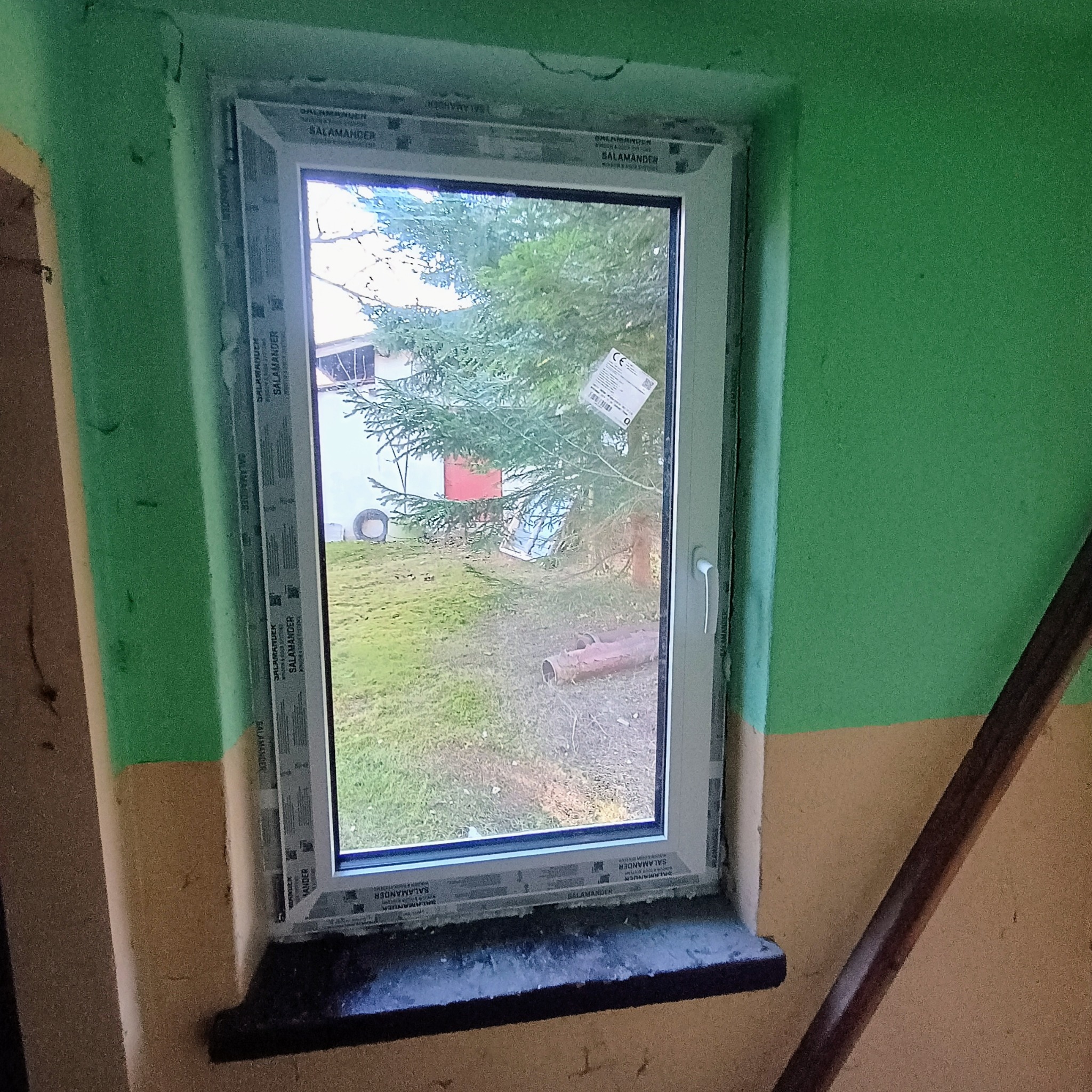 Nowo zamontowane białe okno z widokiem na zewnątrz, z naklejkami producenta na ramie, osadzone w surowym, zielono-beżowym murze.