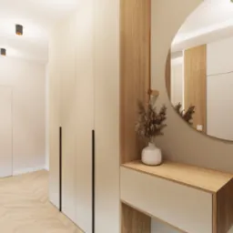 Jasny hol z szafą w zabudowie, okrągłym lustrem i drewnianymi lamelami. Na komodzie wazon z suchymi trawami. Parkiet w jodełkę. Minimalistyczny design.