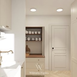 WIKTORIA FIGURA - Jasna kuchnia w stylu klasycznym: białe szafki z mosiężnymi uchwytami, marmurowy blat, złota bateria i drewniane akcenty. Widoczny fragment półki z naczyniami i rośliną.