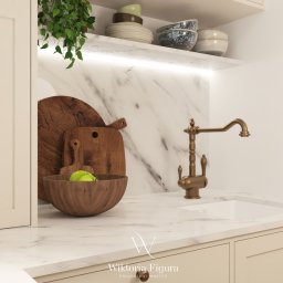 WIKTORIA FIGURA - Kremowa zabudowa kuchenna z marmurowym blatem i złotą armaturą. Drewniane deski i miska z jabłkiem dodają ciepła. Roślina zwisa z półki, oświetlona subtelnym światłem.