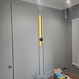 KDW Krzysztof Dusza - Szare panele ścienne z dekoracyjnymi ramkami, nowoczesna lampa ścienna pionowa z czarnymi elementami, biały grzejnik żeberkowy, wystające kable elektryczne i karton z akcesoriami do sprzątania podłóg.