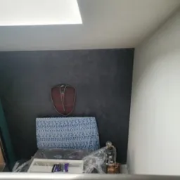 Ściana w odcieniach szarości z dekoracyjną fakturą, ozdobiona zawieszką w kształcie tarczy; widoczny fragment okna z zieloną ścianą oraz meble zabezpieczone folią.