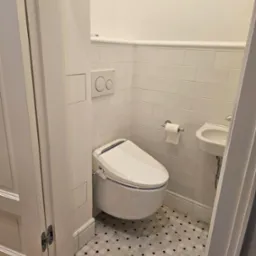 Biała toaleta myjąca z deską bidetową i podwieszana umywalka w małej, wykafelkowanej łazience z wzorzystą podłogą.