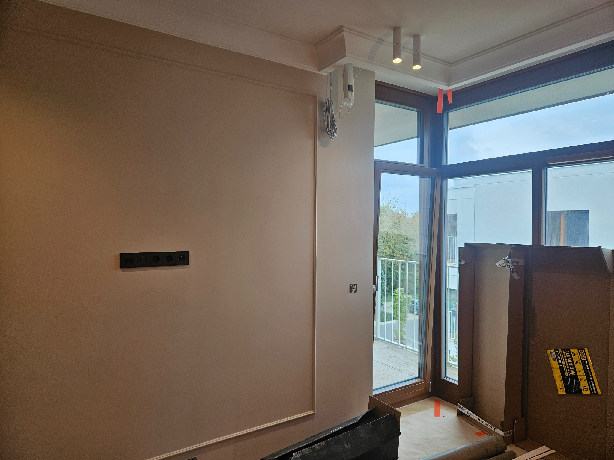 Wnętrze pokoju w trakcie prac wykończeniowych: ściany pomalowane na jasny beż, widoczna instalacja elektryczna, duże okno balkonowe z widokiem na zewnątrz, zabezpieczenia na oknach, kartony...