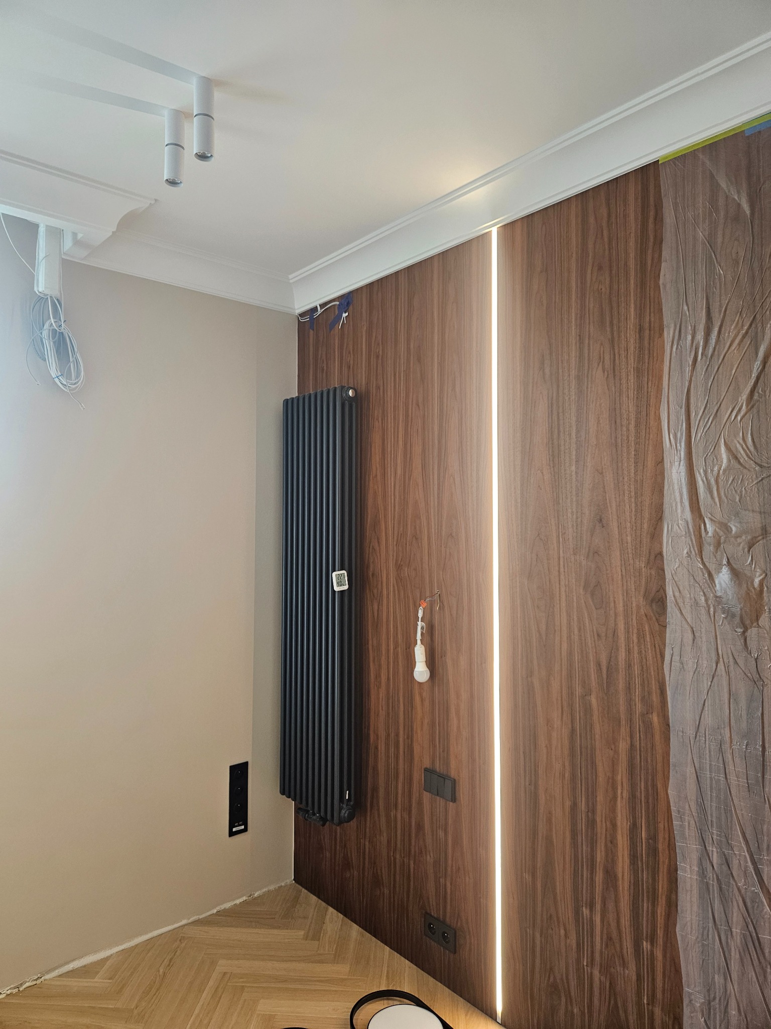 Ściana z paneli drewnopodobnych z podświetleniem LED, czarny grzejnik żeberkowy, beżowa ściana, listwa przypodłogowa w trakcie malowania, zabezpieczona folią część ściany.