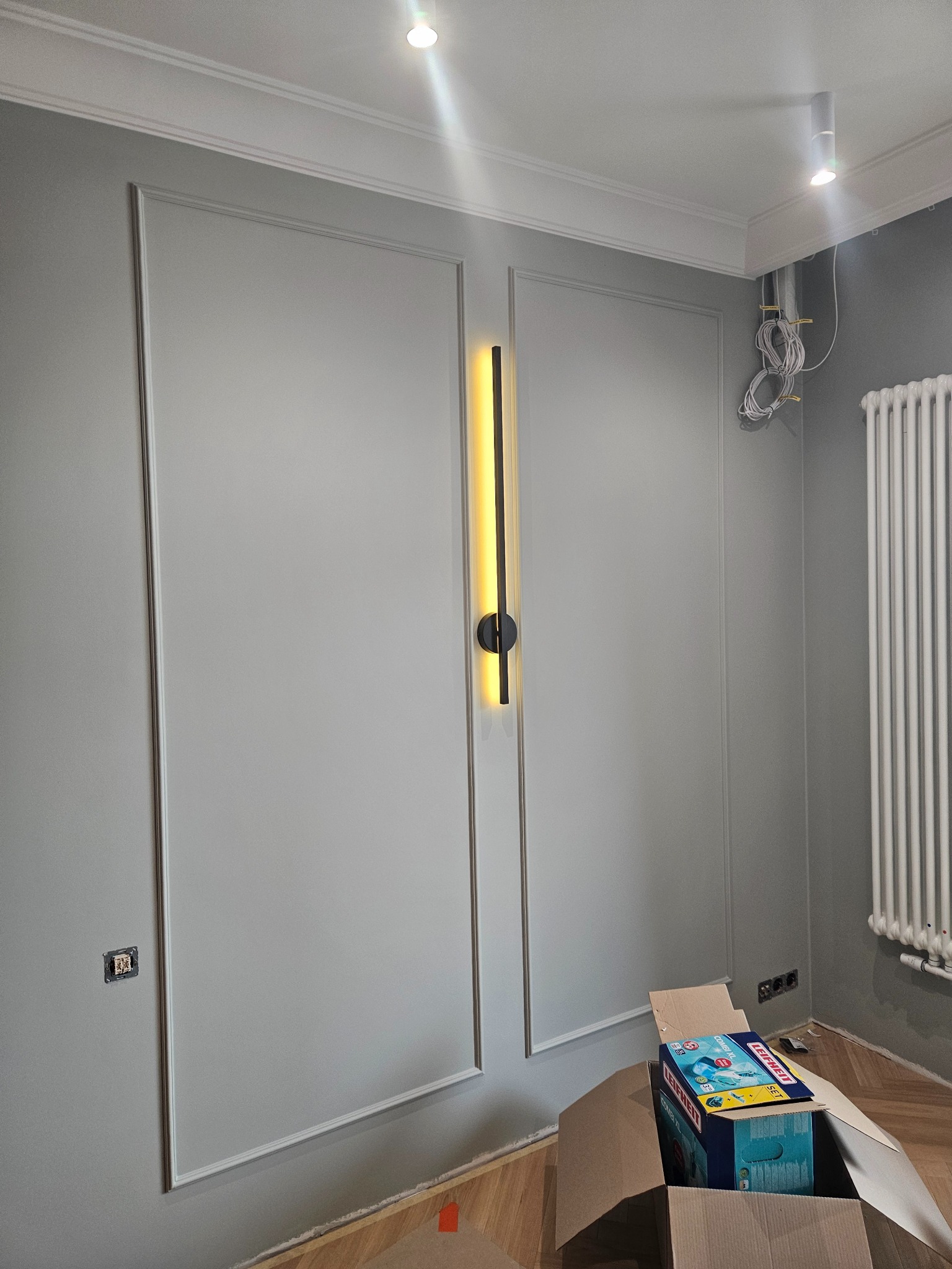 Szare panele ścienne z dekoracyjnymi ramkami, nowoczesna lampa ścienna pionowa z czarnymi elementami, biały grzejnik żeberkowy, wystające kable elektryczne i karton z akcesoriami do sprzątania podłóg.