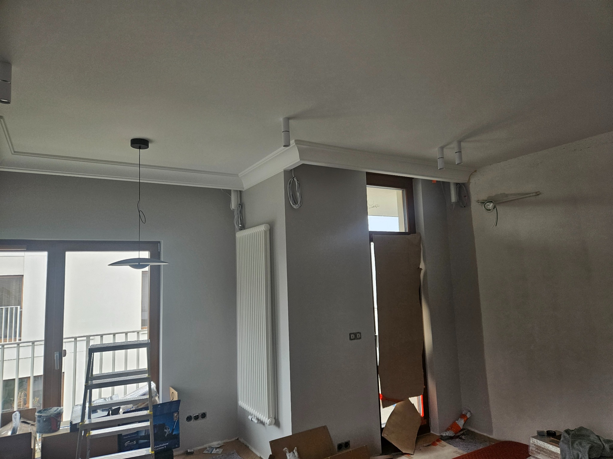 Wnętrze w trakcie prac wykończeniowych: szare ściany, zamontowany grzejnik, listwy przysufitowe z oświetleniem punktowym, drabina oparta o okno balkonowe, widoczne puszki elektryczne i przewody...