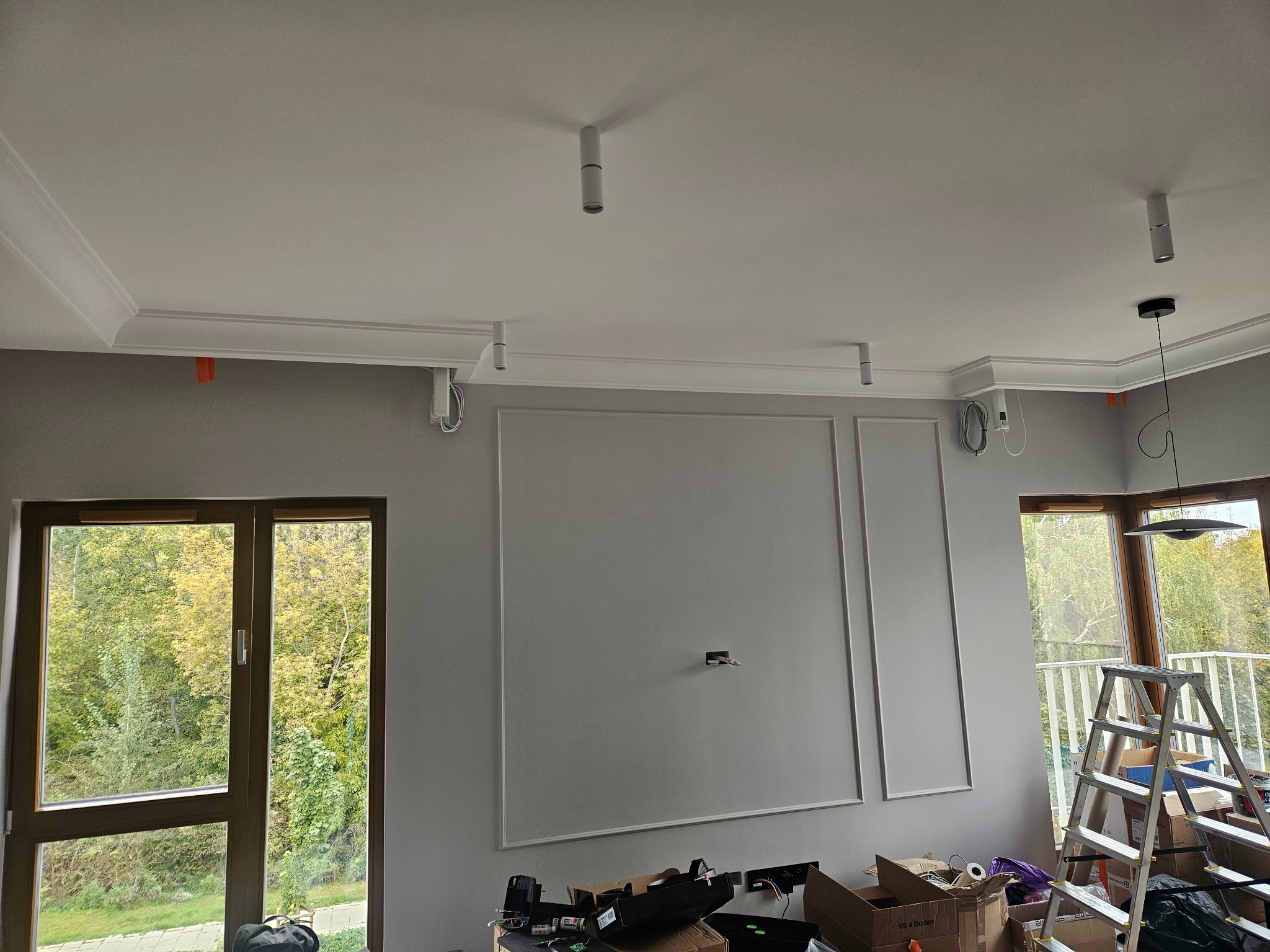 Szare wnętrze w trakcie wykańczania z dekoracyjnymi listwami ściennymi, sztukaterią sufitową, zamontowanymi reflektorami punktowymi i widoczną instalacją elektryczną, skrzynkami i drabiną w tle.