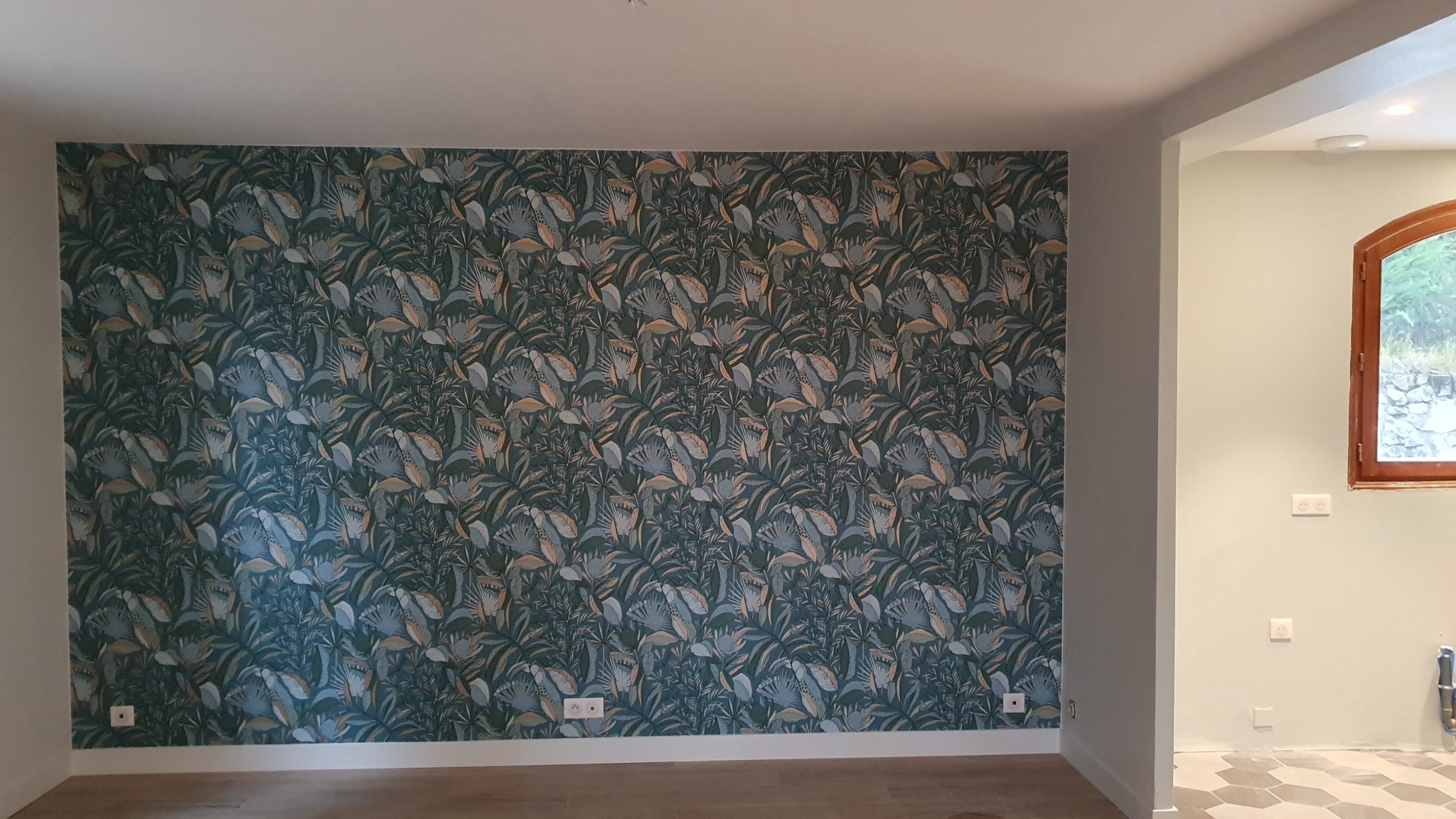 Ściana z wzorzystą, zielono-niebieską tapetą w motywy roślinne, widok na fragment pomieszczenia z drewnianą ramą okienną i jasnymi ścianami.