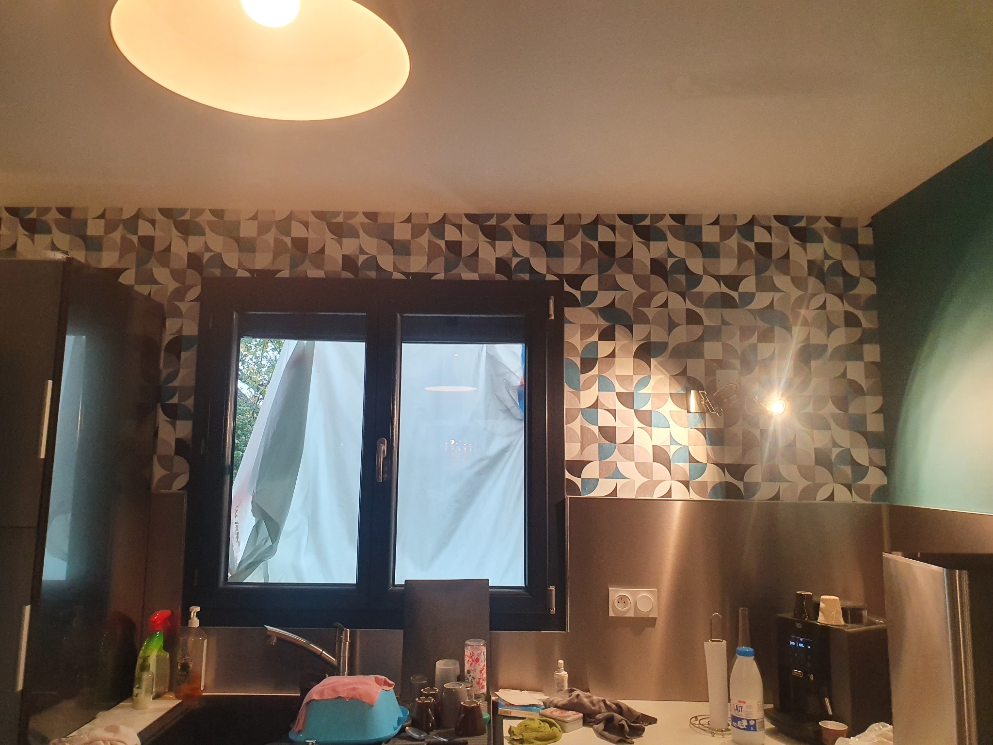 Kuchnia po odświeżeniu: wzorzysta tapeta w odcieniach szarości i błękitu nad metalowym panelem, widok na okno z zasłoniętą szybą, oświetlenie sufitowe i punktowe, fragment wyposażenia kuchennego...