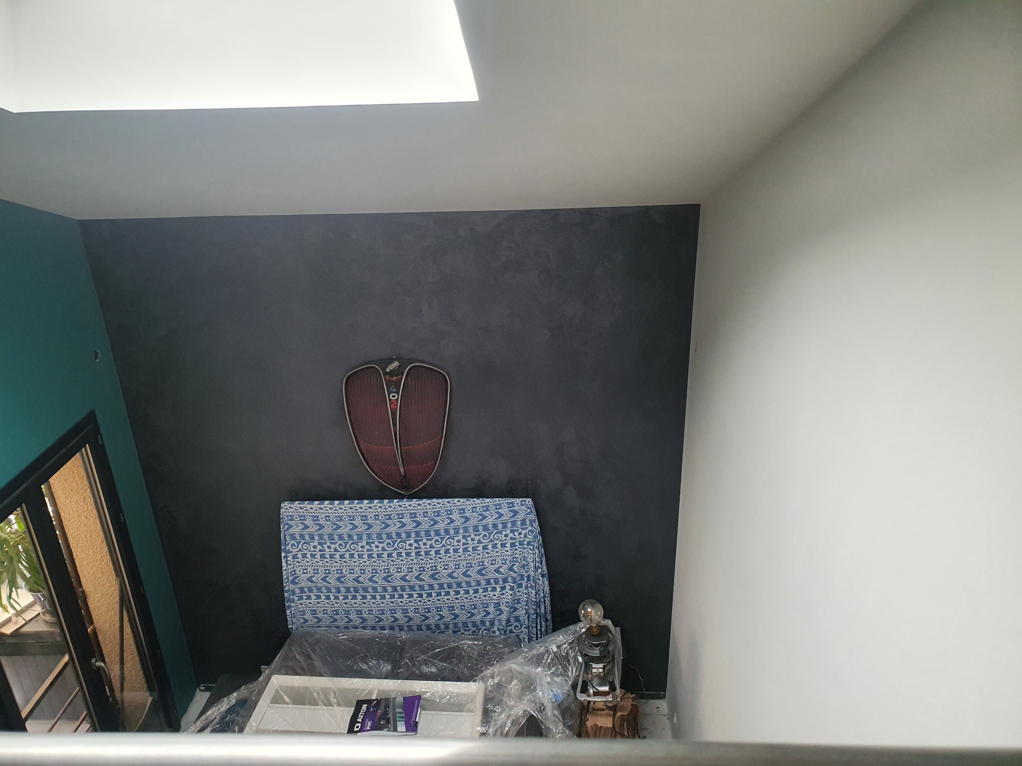 Ściana w odcieniach szarości z dekoracyjną fakturą, ozdobiona zawieszką w kształcie tarczy; widoczny fragment okna z zieloną ścianą oraz meble zabezpieczone folią.