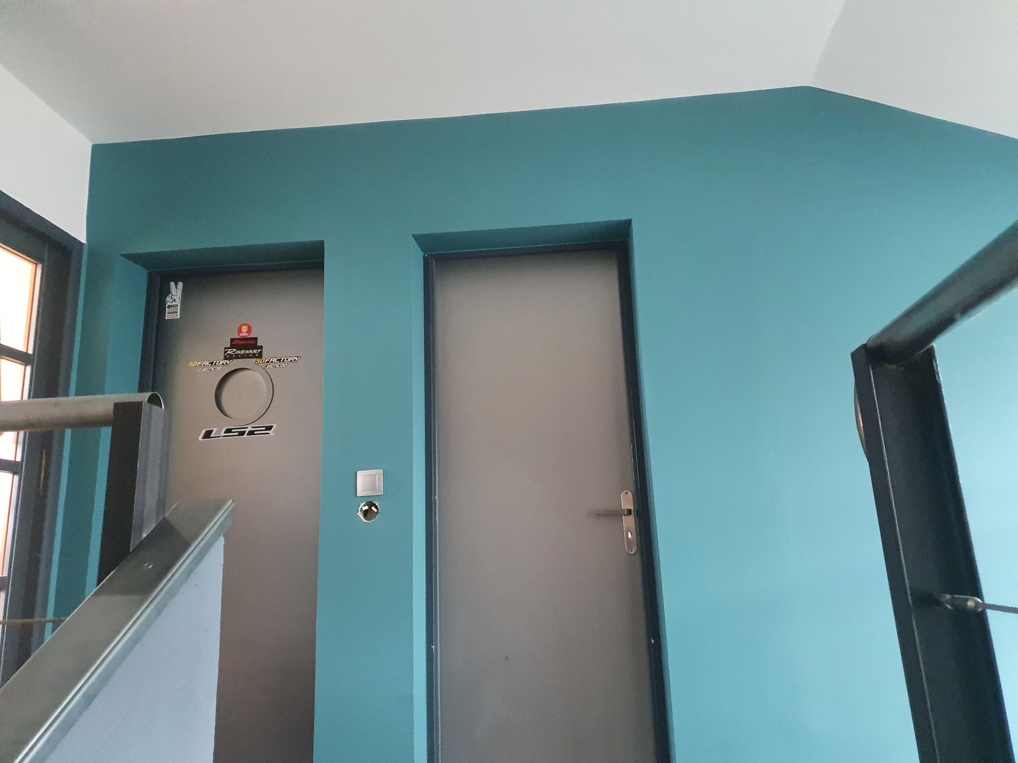Dwa szare, metalowe drzwi w turkusowej ścianie, z widoczną metalową barierką schodów w tle.