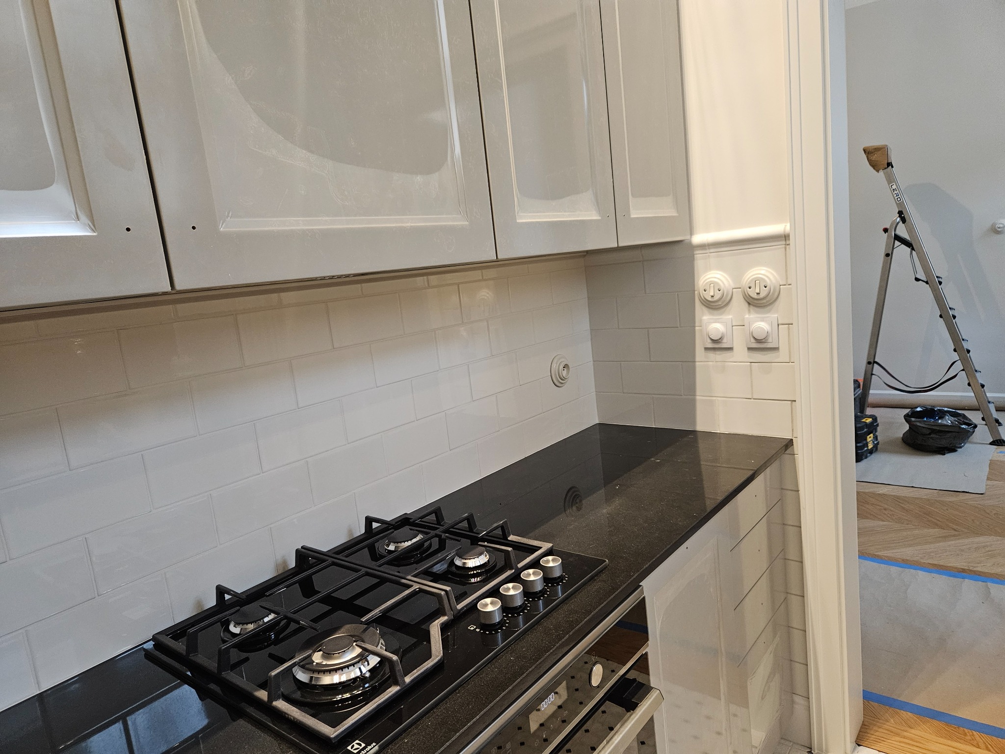 Nowoczesna kuchnia po remoncie: szare szafki, białe płytki ścienne, czarny blat, płyta gazowa Electrolux i odnowione gniazdka z przełącznikami w stylu retro, drabina oparta o ścianę sugeruje...