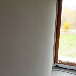 MG LuxTynk - Świeżo otynkowana ściana wewnętrzna z widocznym oknem i fragmentem podłogi, szare wykończenie, widoczny kabel przy podłodze.