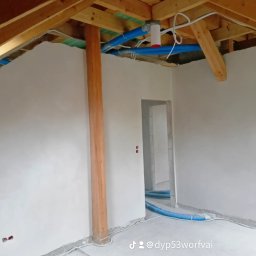MG LuxTynk - Wnętrze w trakcie prac wykończeniowych, świeżo otynkowane ściany, widoczne drewniane elementy konstrukcyjne dachu, niebieskie rury wentylacyjne pod sufitem i na podłodze, przygotowane do dalszych...
