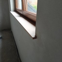 MG LuxTynk - Surowe ściany z oknem z drewnianą ramą i parapetem w trakcie prac wykończeniowych, widoczne kable przy podłodze.