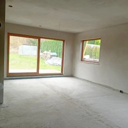 MG LuxTynk - Surowe wnętrze z dużym oknem przesuwnym i dwoma mniejszymi, ściany po nałożeniu gładzi, w tle widok na ogród z tujami i blaszakiem, na podłodze widoczny walec folii ochronnej.