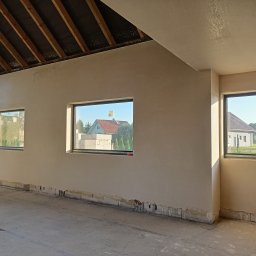 MG LuxTynk - Surowe wnętrze z oknami po nałożeniu tynku, widoczne niedoskonałości przy podłodze i fragment drewnianej konstrukcji dachu.