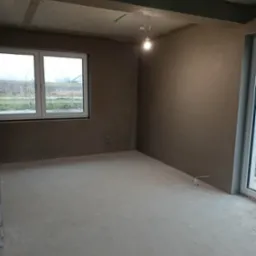 Surowe wnętrze po nałożeniu gładzi gipsowej na ściany, widoczne przyłącza instalacji, surowy betonowy strop i prowizoryczne oświetlenie żarówką