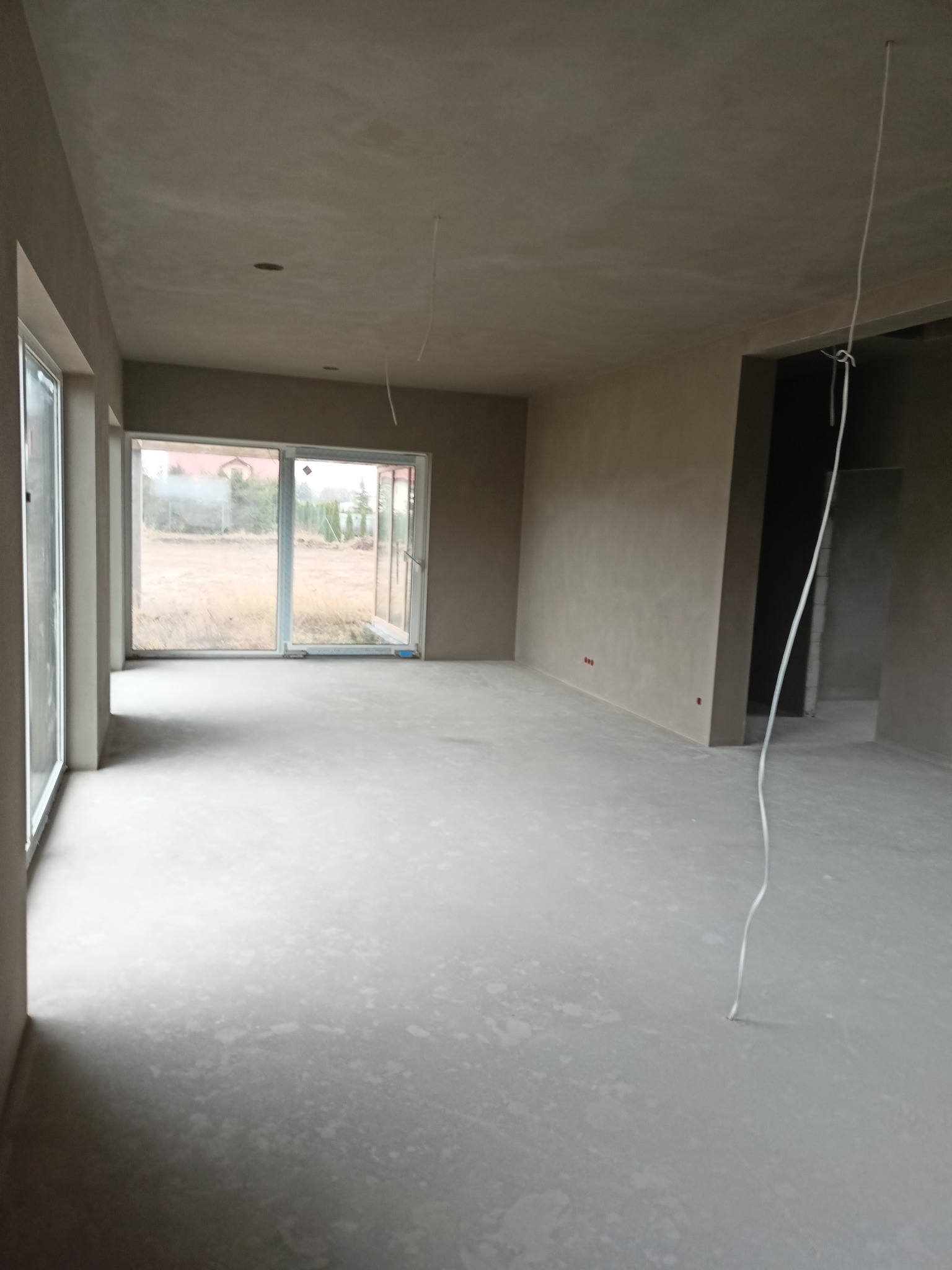 Surowe wnętrze domu w Kętrzynie po etapie prac wykończeniowych: ściany pokryte szarym tynkiem, wylewka na podłodze, widoczne przewody zwisające z sufitu i okno z widokiem na zewnątrz.