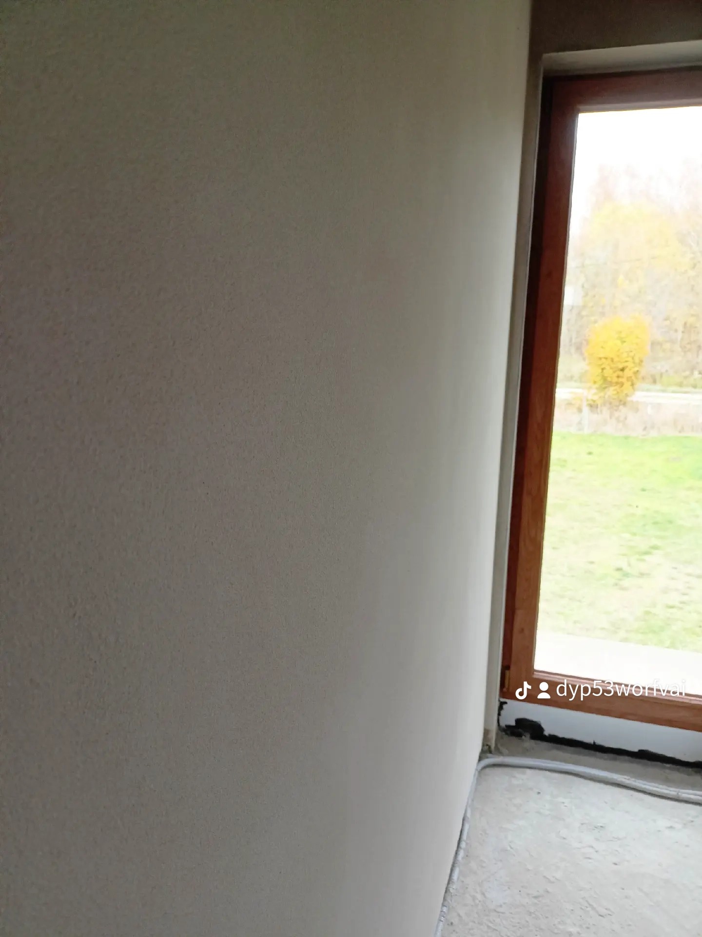 Świeżo otynkowana ściana wewnętrzna z widocznym oknem i fragmentem podłogi, szare wykończenie, widoczny kabel przy podłodze.