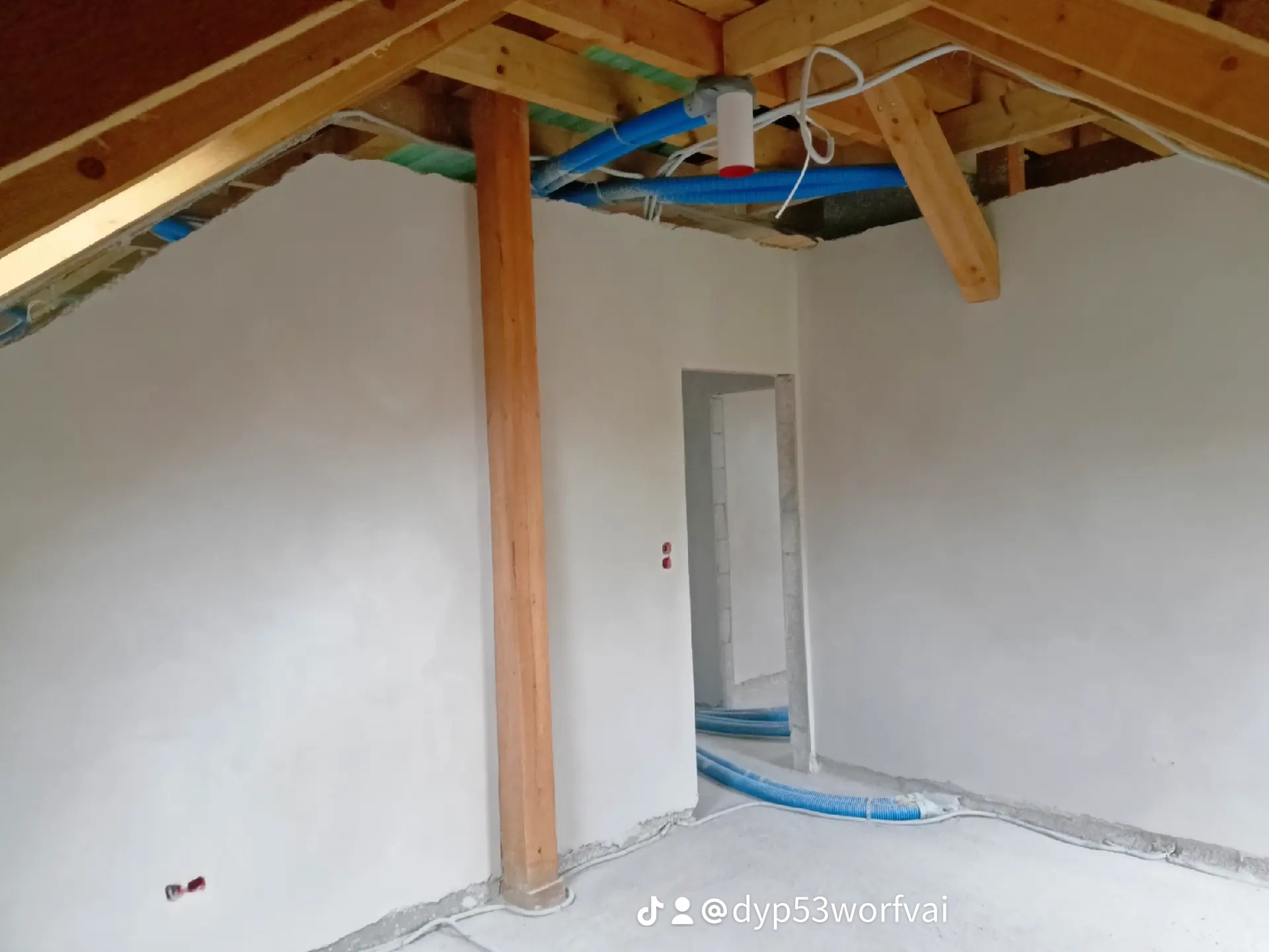 Wnętrze w trakcie prac wykończeniowych, świeżo otynkowane ściany, widoczne drewniane elementy konstrukcyjne dachu, niebieskie rury wentylacyjne pod sufitem i na podłodze, przygotowane do dalszych...